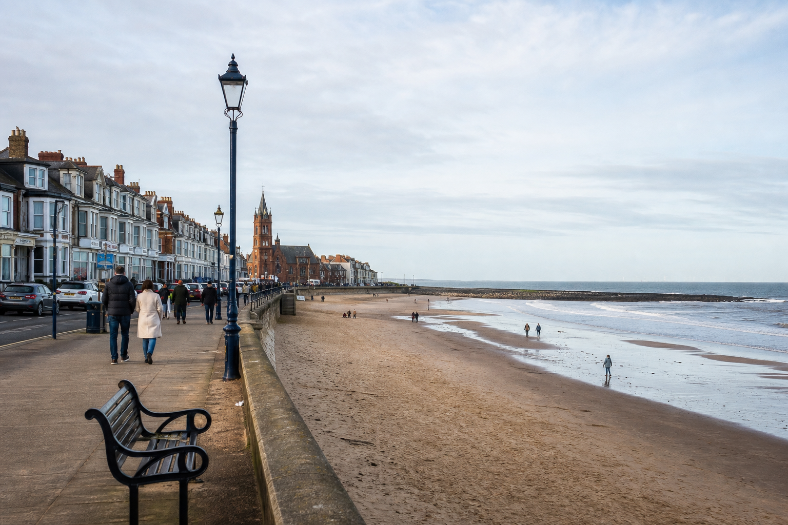 A coastal promenade with people walking, adjacent to sandy beach and seaside town buildings.