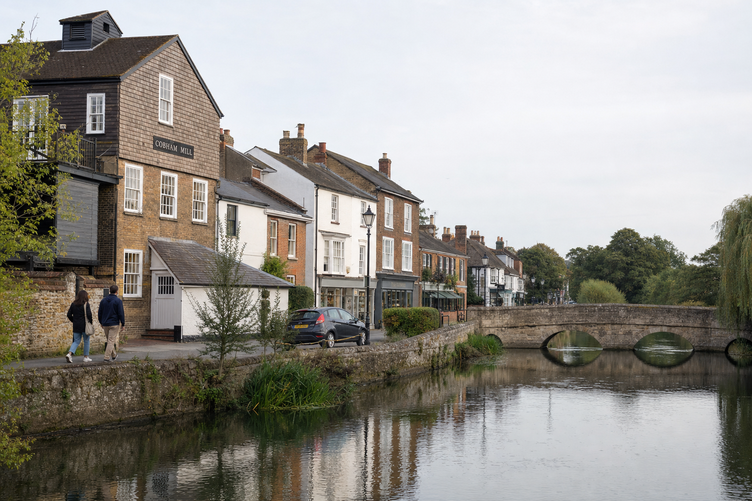 A couple strolls along a riverside path beside Cobham Mill and historic buildings in a quaint English village setting.