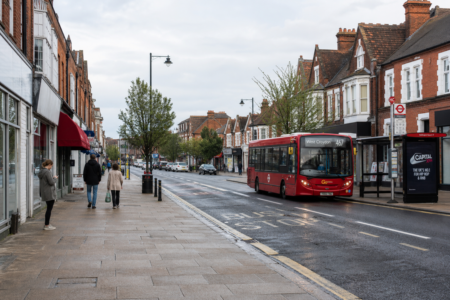 Red bus on a quiet street with people walking, passing shops and bus stop signage, under an overcast sky.