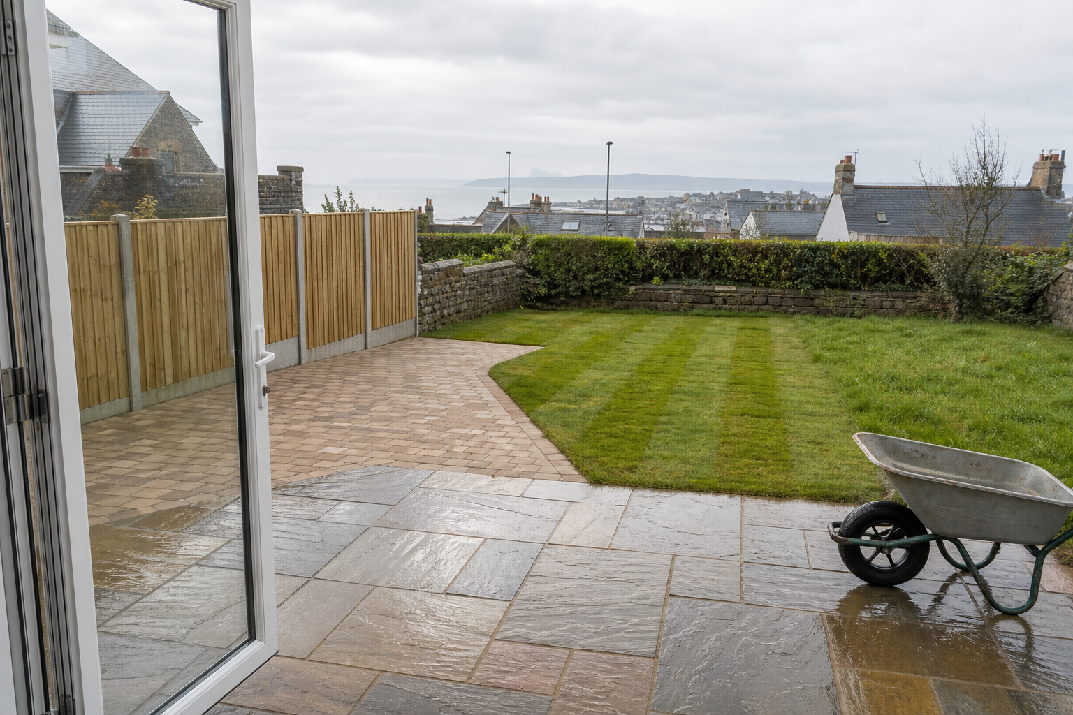 A neatly mowed garden with a wheelbarrow on a wet patio, overlooking houses and the sea in the distance.