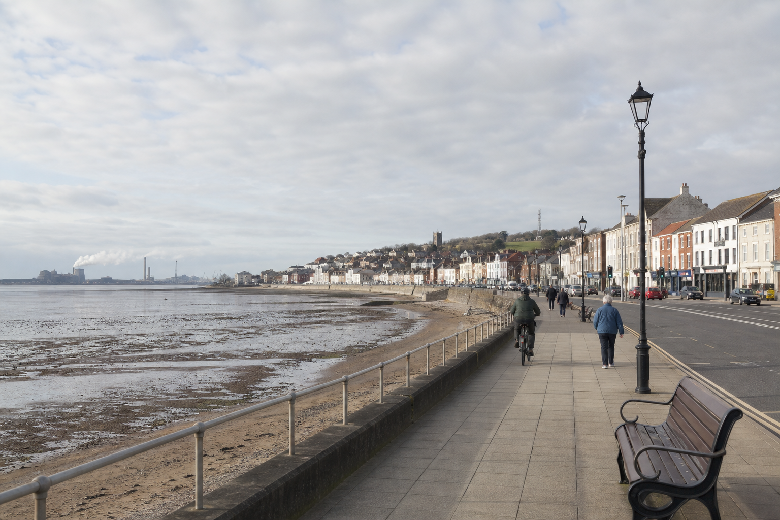 Seaside promenade with people walking and cycling, lined with buildings and overlooking a bay at low tide under a cloudy sky.