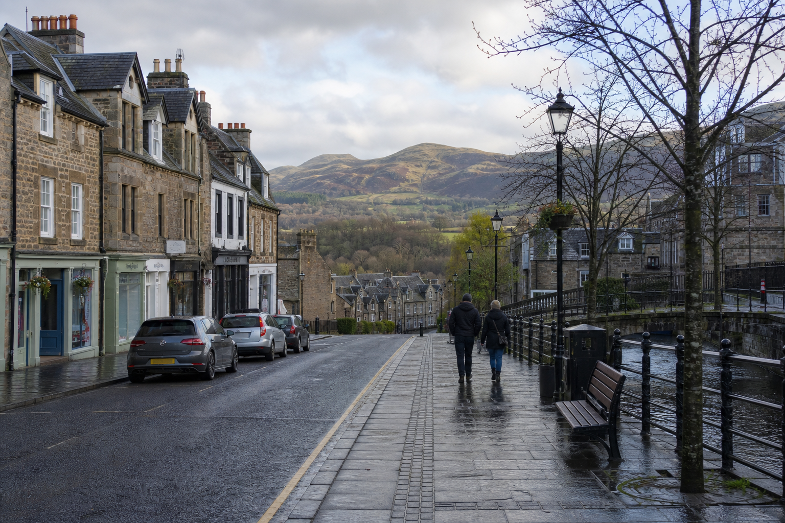 A couple walks along a wet street in a historic town with distant hills visible under a cloudy sky.