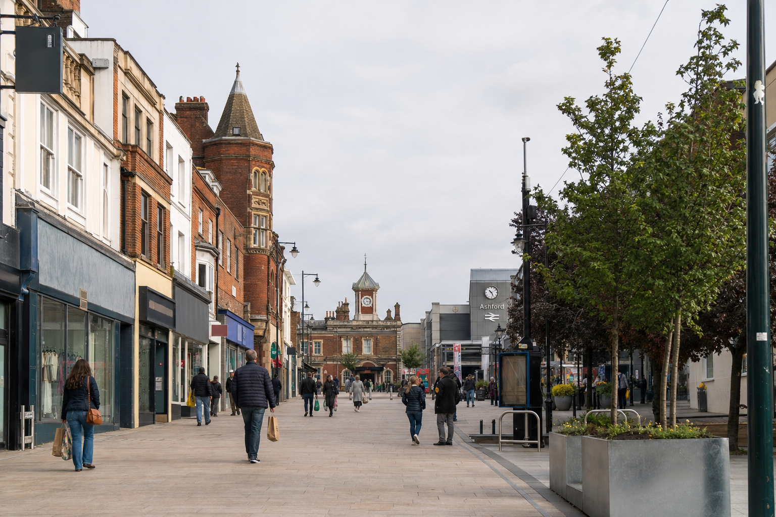 Pedestrians walk along a high street lined with shops and trees, with a clock tower visible in the background.