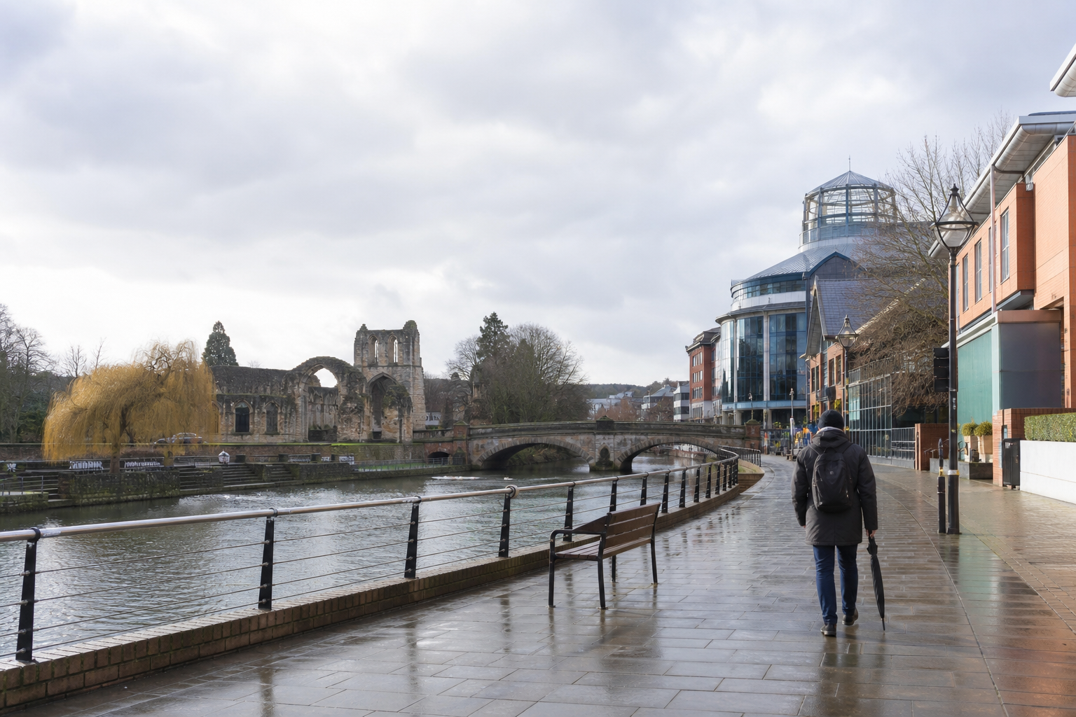Person walking along a riverside path with historic ruins and modern buildings in the background.