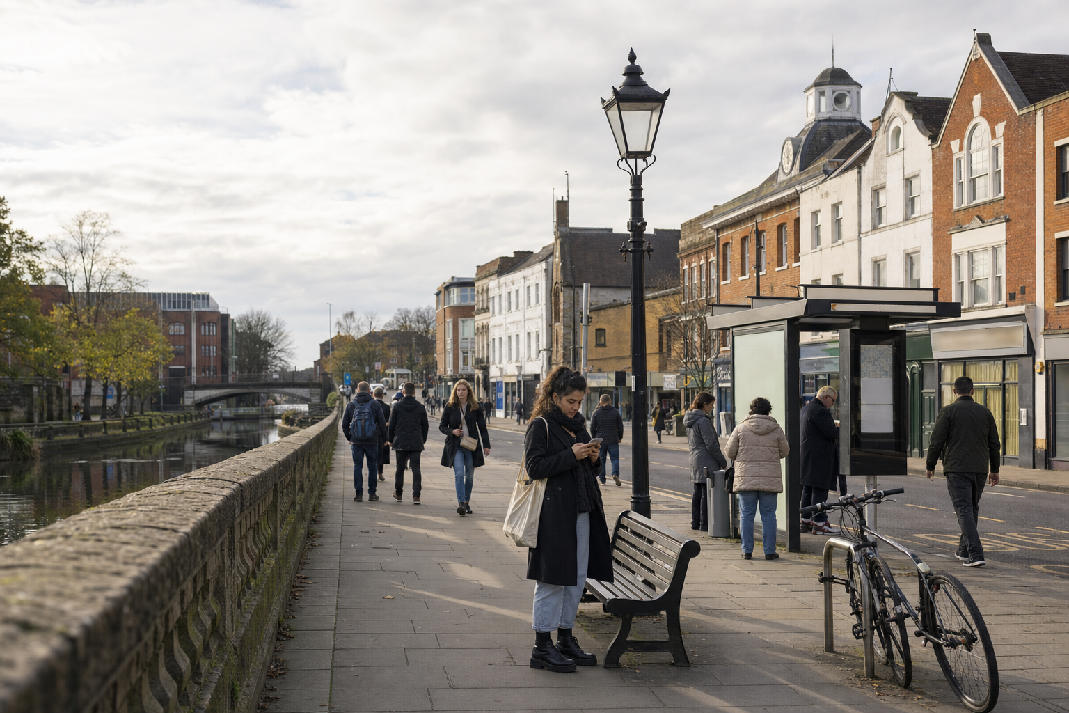 People walking along a riverside pavement with benches, a bicycle, and a bus shelter under a cloudy sky.