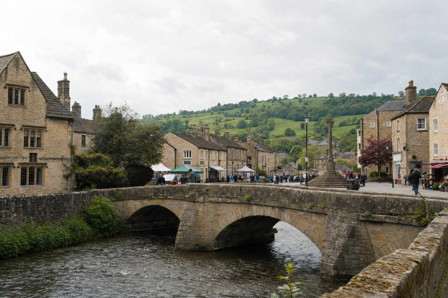 Stone bridge over a river in a quaint village with historic buildings and lush green hills in the background.