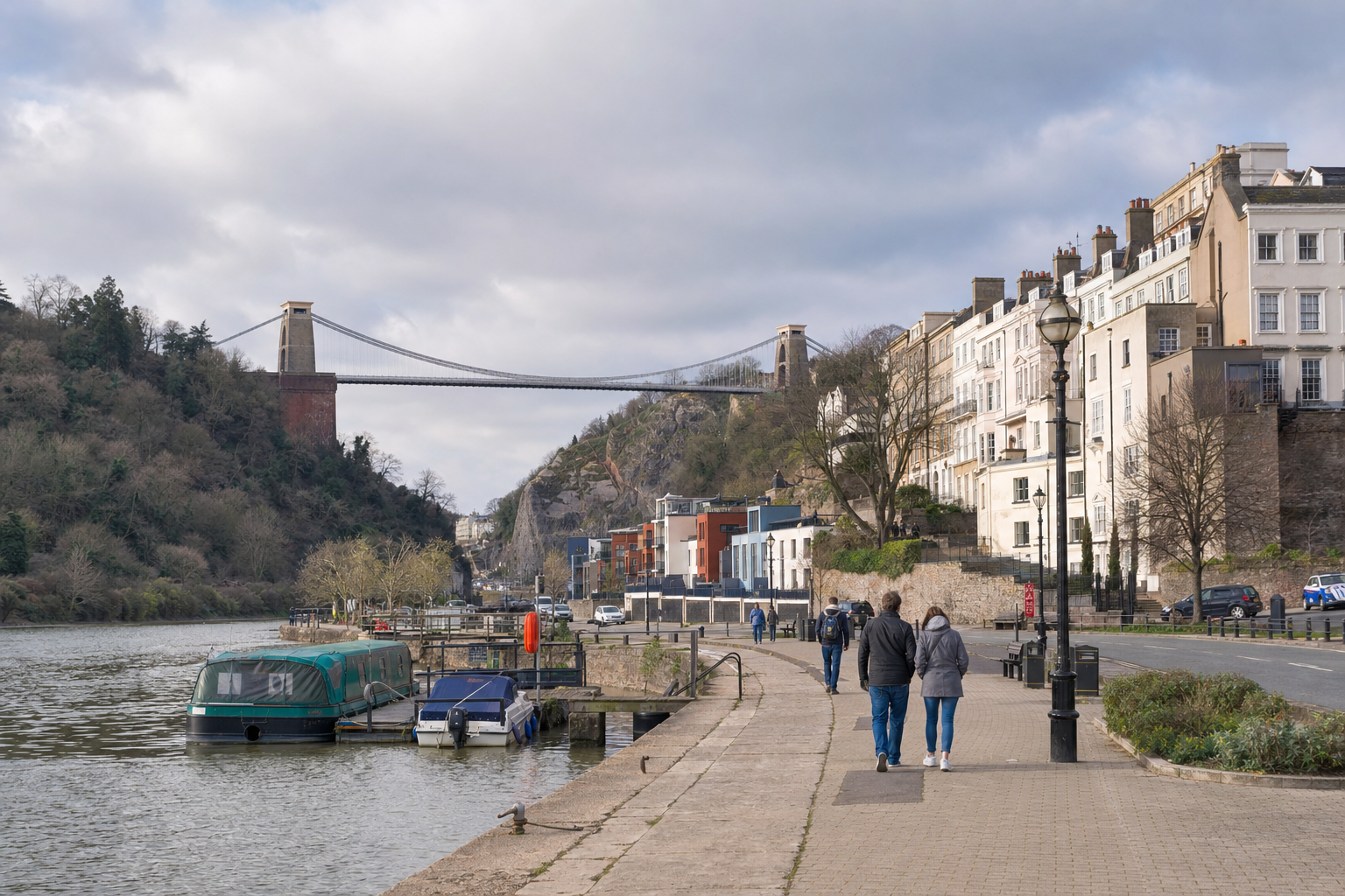 People walk along a riverside path with the Clifton Suspension Bridge in the background under a cloudy sky.