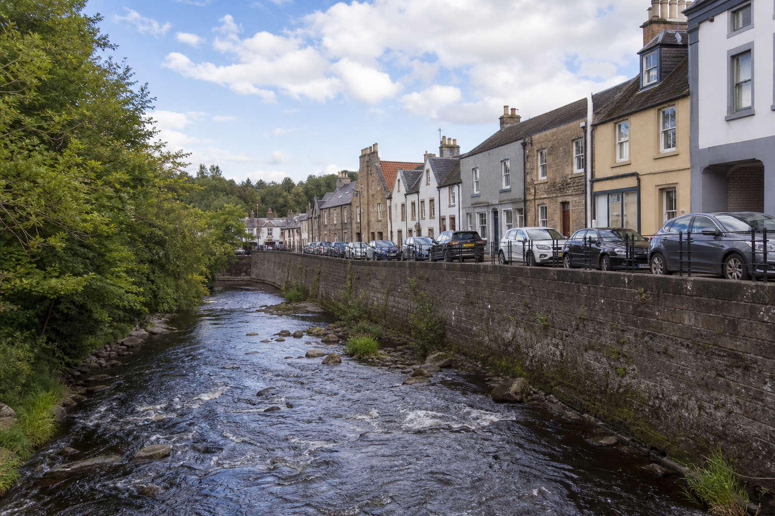 A tranquil river flows between leafy trees and a row of parked cars beside charming stone buildings.