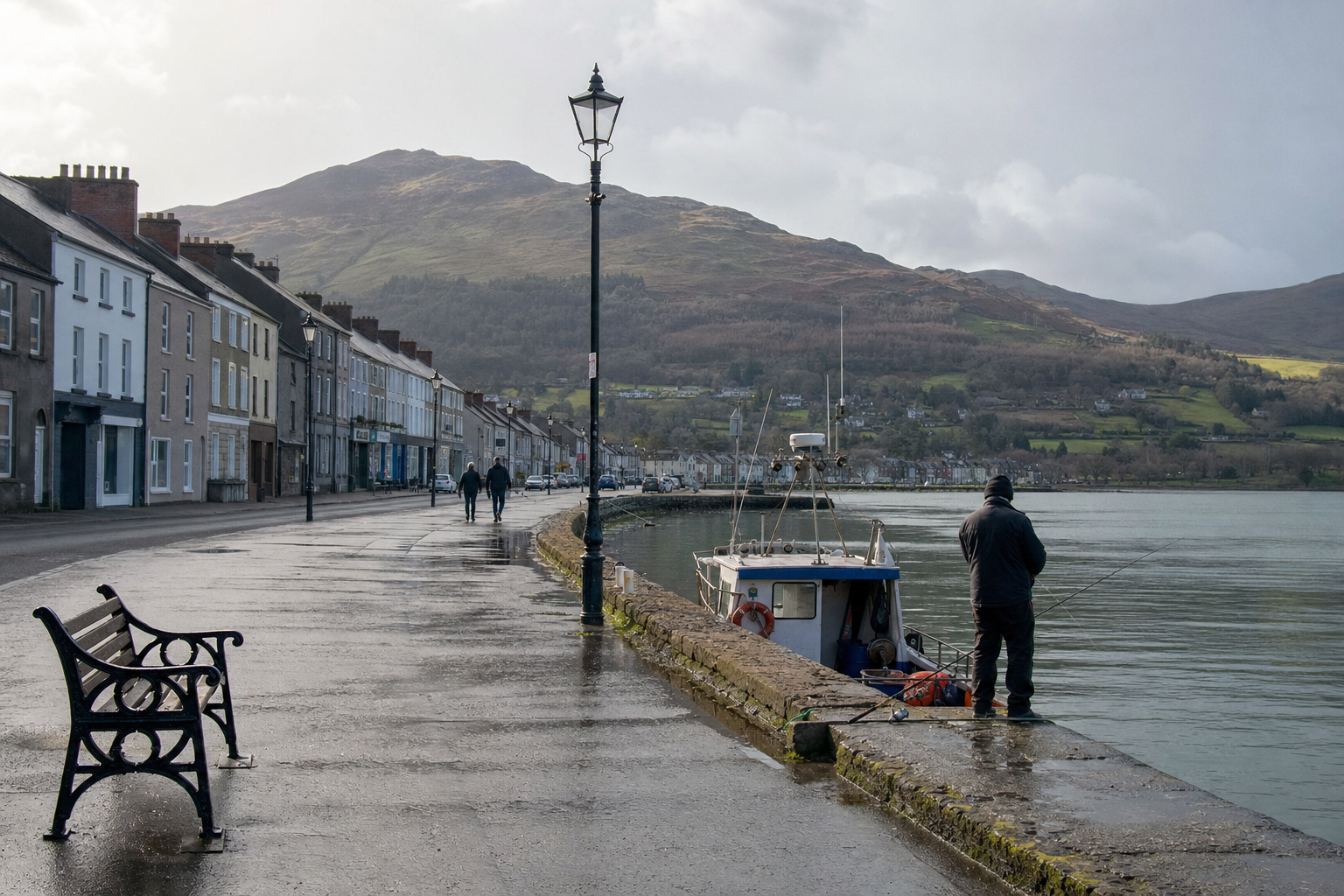 A person fishing near a boat on a wet promenade with a row of houses and a hill in the background.