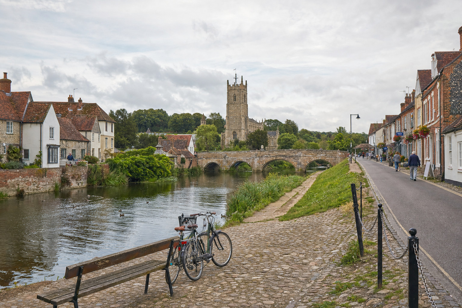Historic stone bridge and church tower overlooking a riverside village street with houses and a bench with bicycles.