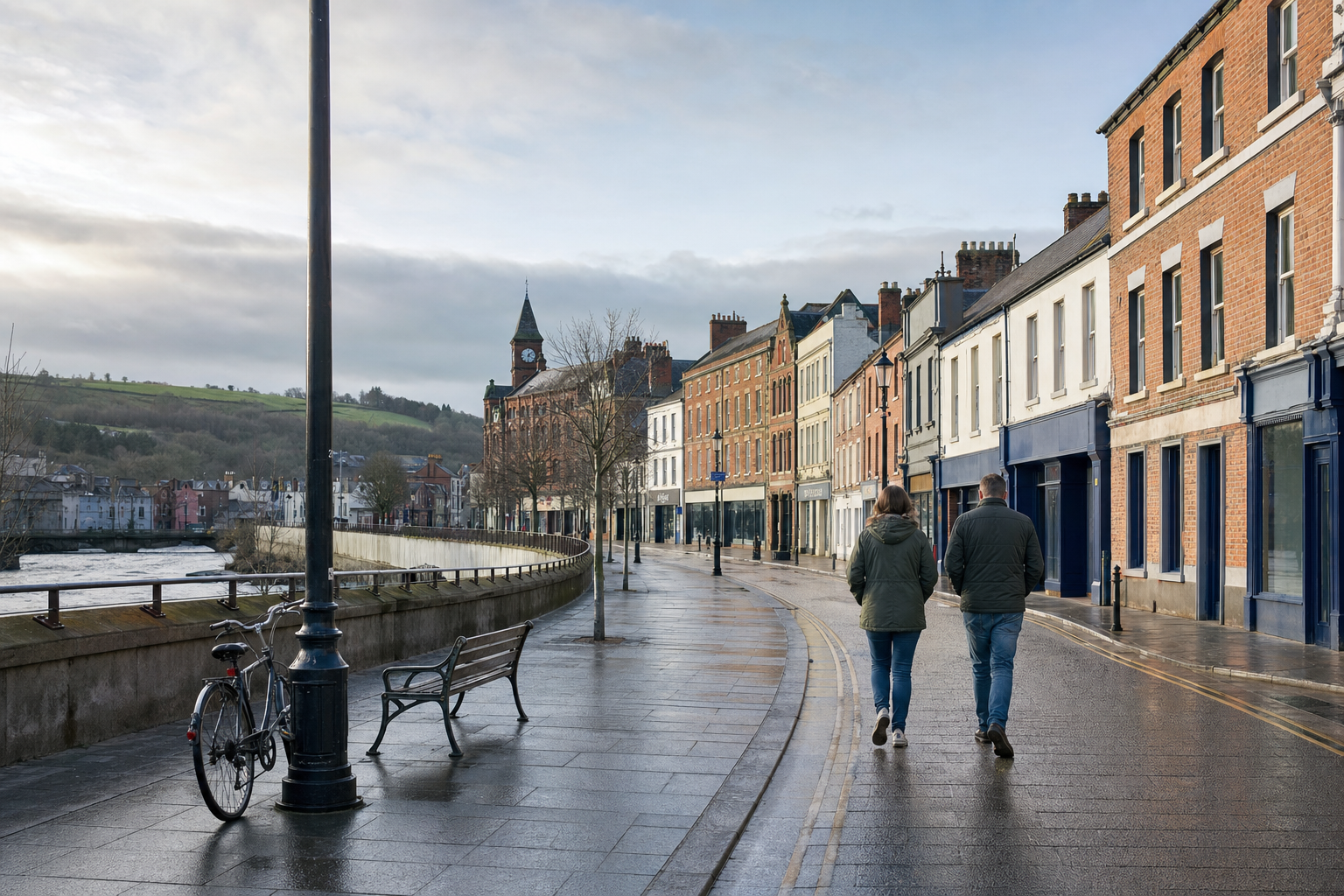 A couple walks along a riverside pavement beside colourful historic buildings on a cloudy day.