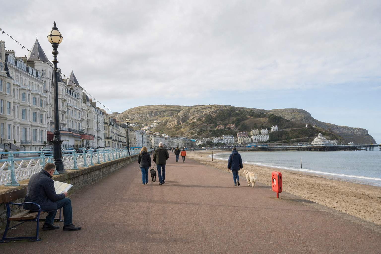 People walking along a seaside promenade with a hillside and pier in the background.