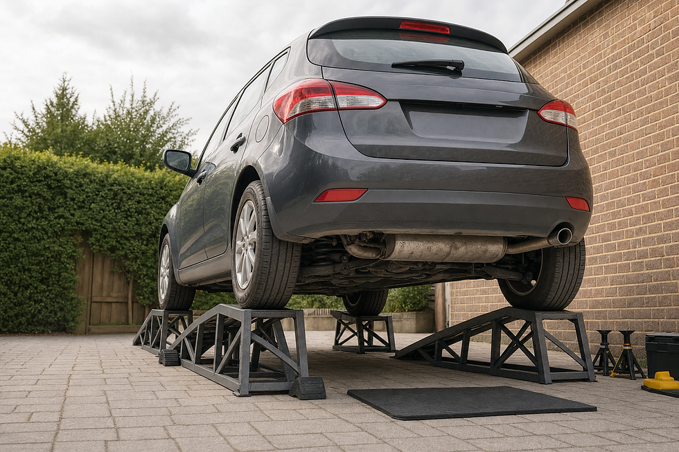 A grey car is elevated on ramps in a driveway next to a brick wall, with tools and jack stands nearby.