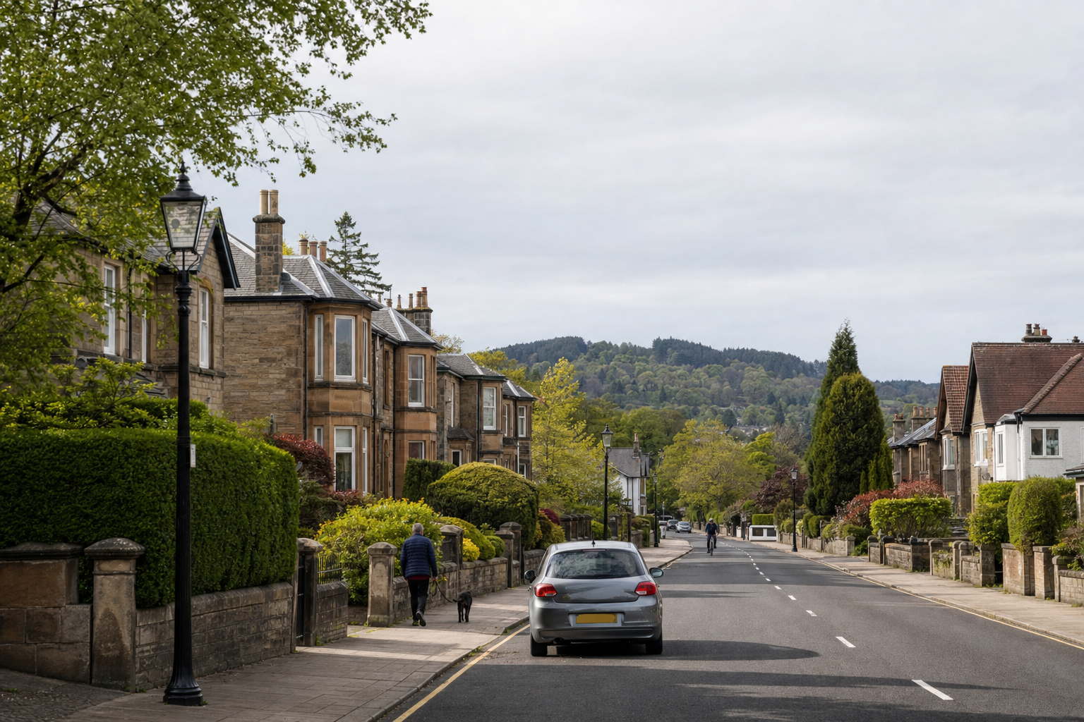 Residential street with stone terraced houses, a parked car, and a person walking a dog on a tree-lined pavement.