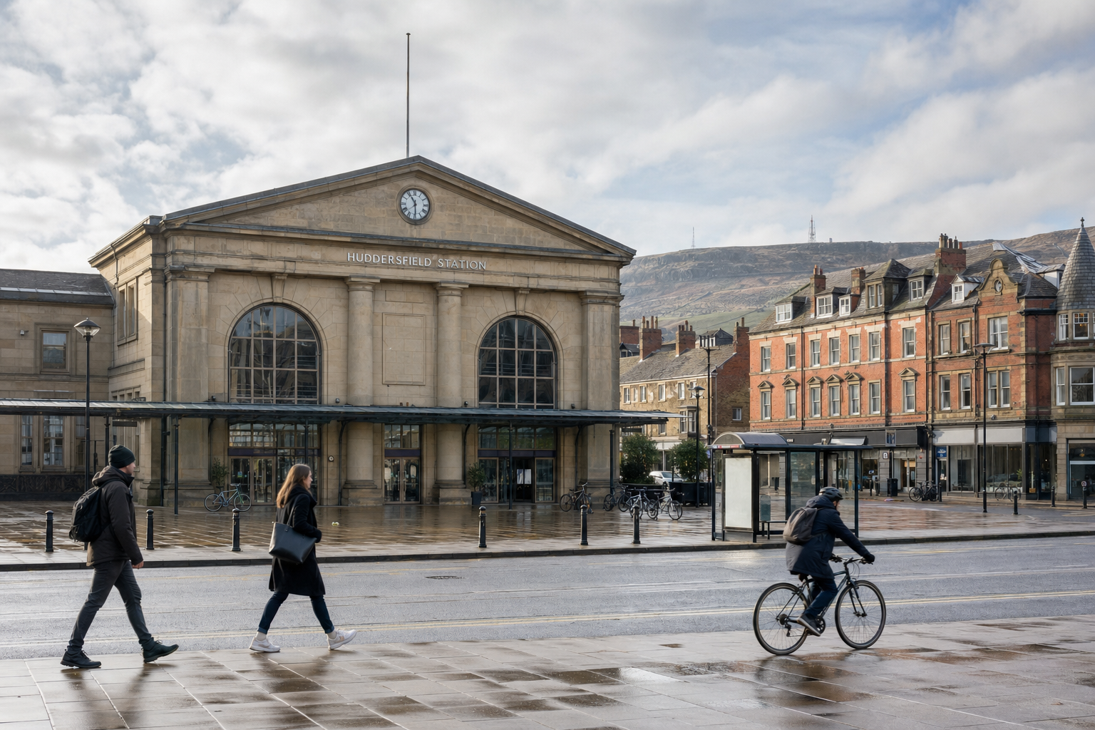 People walk and cycle in front of the historic Huddersfield railway station on a cloudy day.