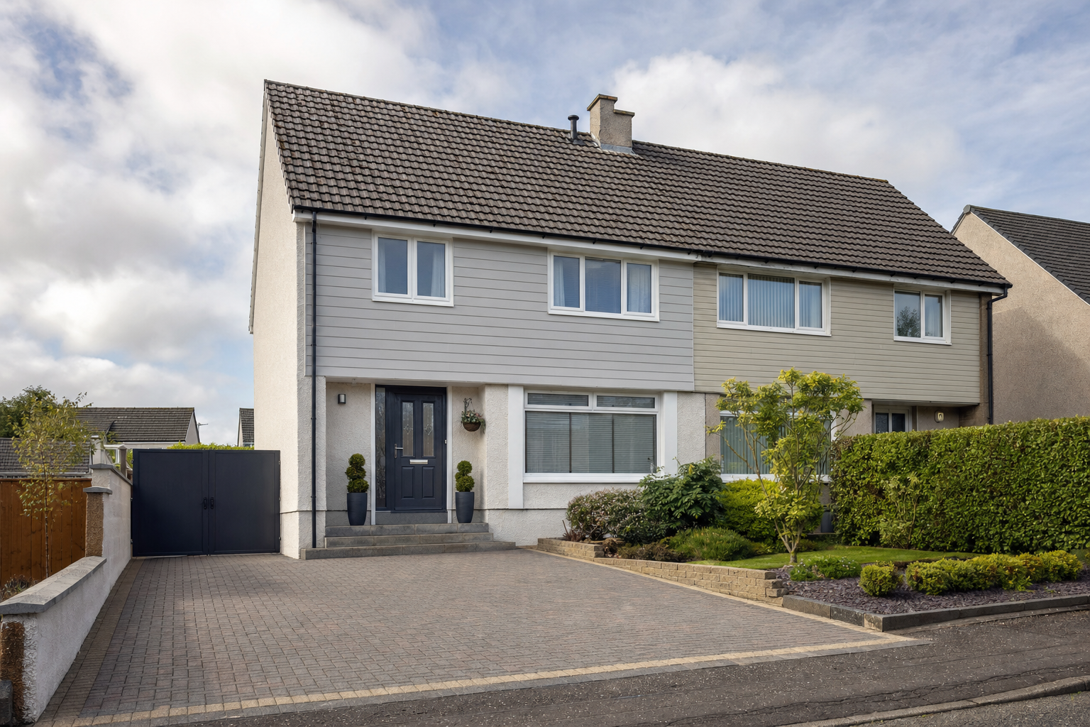 A semi-detached house with a grey facade, neatly trimmed bushes, and a paved driveway under a cloudy sky.