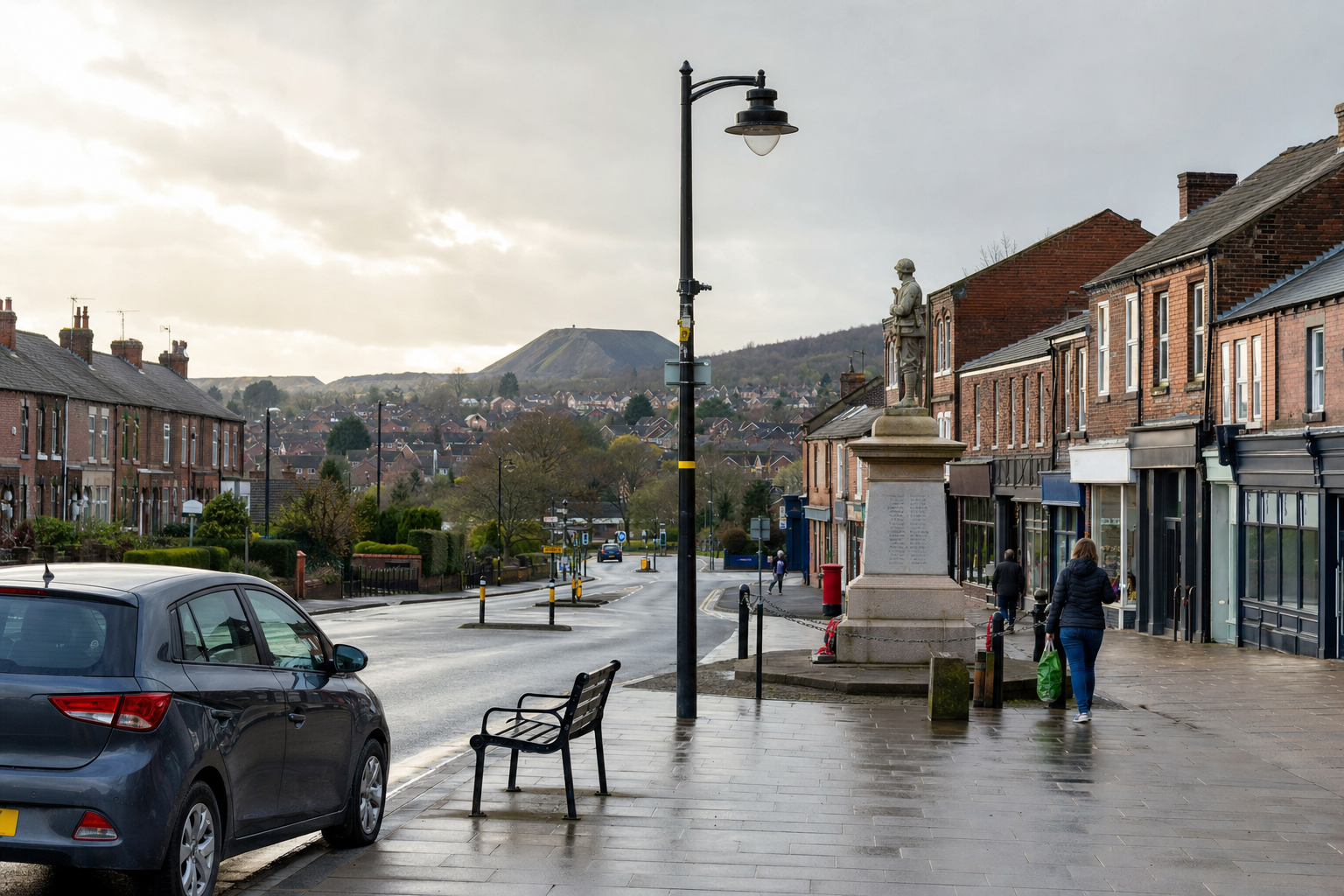 A quiet street scene with a war memorial statue, parked car, brick buildings, and a distant hill under a cloudy sky.