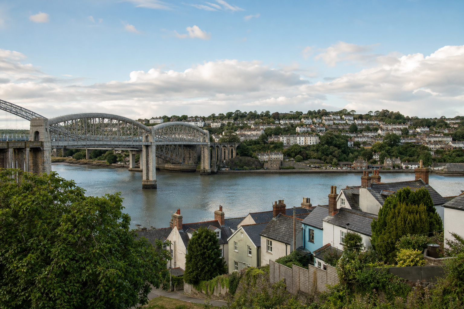 A scenic view of a bridge over a river with houses and trees in the foreground and hillside homes in the background.