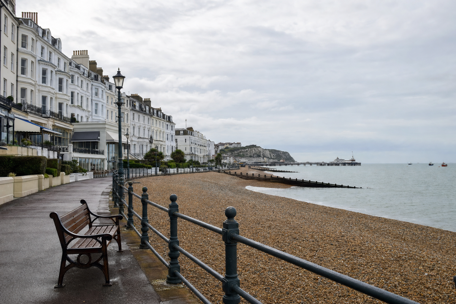 A seaside promenade with benches and historic white buildings along a pebble beach view.