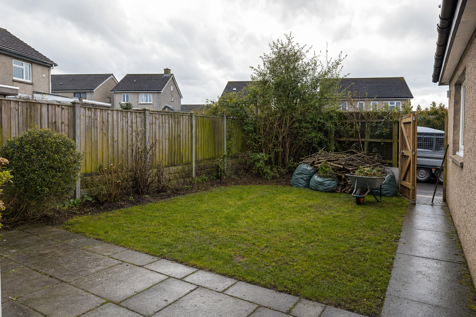 A small, tidy garden with a pile of branches and a wheelbarrow next to a wooden fence on a cloudy day.