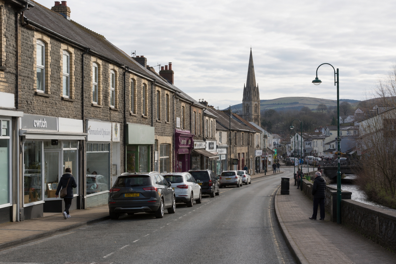 A quiet British high street with parked cars, shops, and a tall church spire in the distance under a cloudy sky.