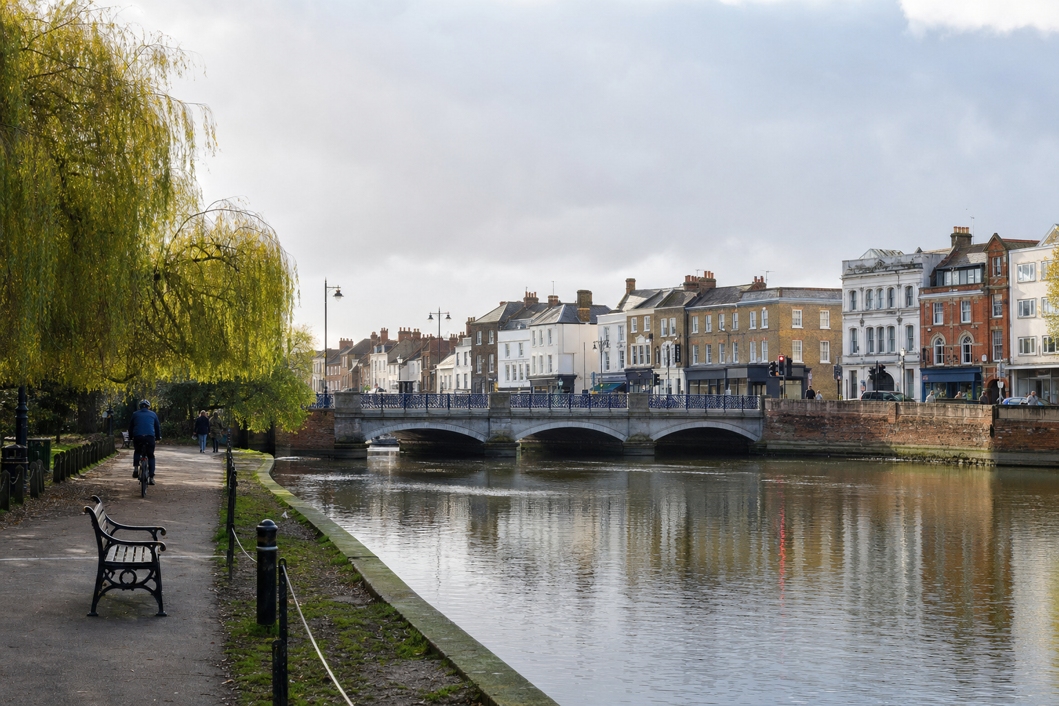 Cyclist on a riverside path with a view of a bridge and classic townhouses under a cloudy sky.