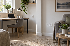 A cosy home office setup with a laptop on a wooden desk, grey chair, and plants, beside a comfy sofa and coffee table.