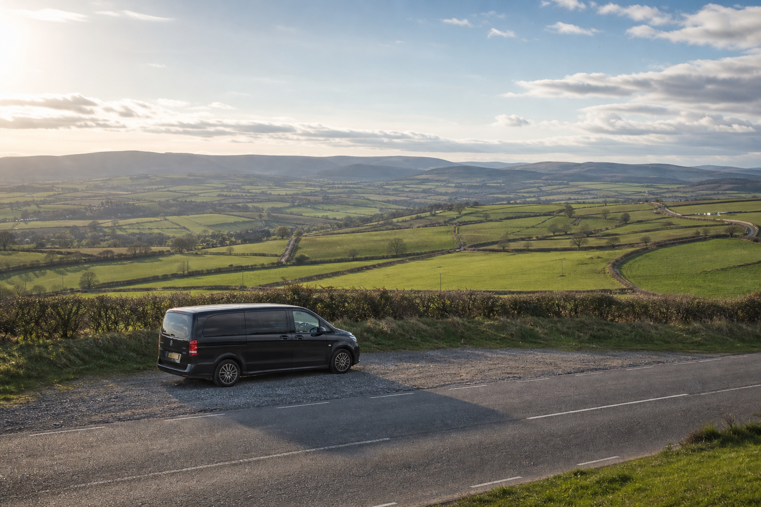 A black van is parked beside a country road overlooking expansive green fields and distant hills under a partly cloudy sky.