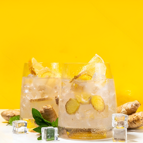 Two glasses of ginger lemonade with ice cubes and lemon slices on a white table. Bright yellow background enhances the refreshing mood.