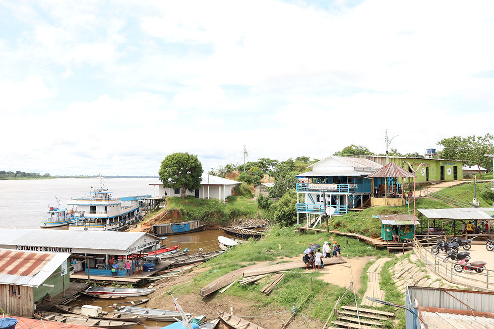 Riverside scene with moored boats, blue and green buildings. People walk on a dirt path under a cloudy sky.