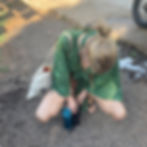 A person in a green patterned outfit crouches over a black puppy on a gravel path. They carry a tote with a sun symbol and a blue bottle.