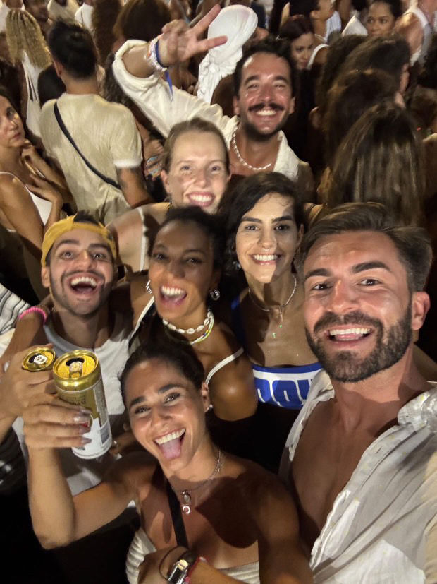 A group of smiling people at a party, holding drinks. They're wearing casual summer outfits. The background shows a lively crowd.