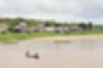 Two people in a small boat on a river, near a village with colorful houses and greenery.
