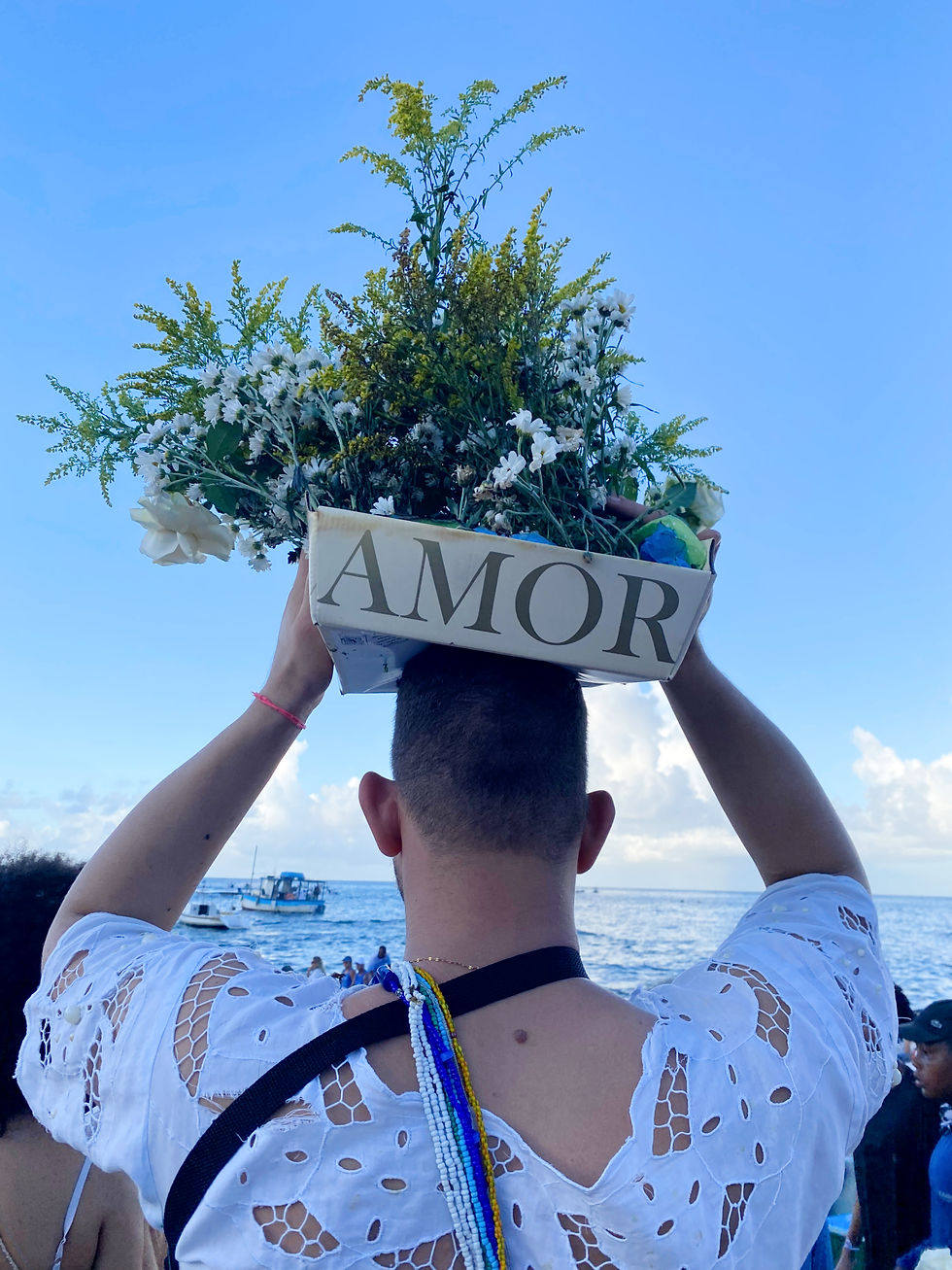 Person in white holds a box with "AMOR" and flowers above their head at the beach, under a blue sky. People and boats in the background.