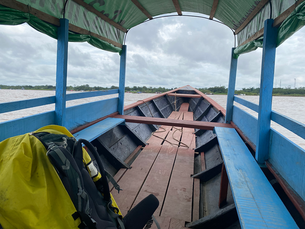 Inside a wooden boat with blue and red accents, a yellow rain cover on a backpack. Overcast sky, river, and green landscape visible.