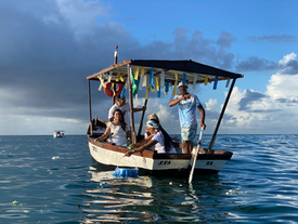 A small boat with three people on calm sea, under a blue sky. The boat is decorated with colorful streamers; one person rows.