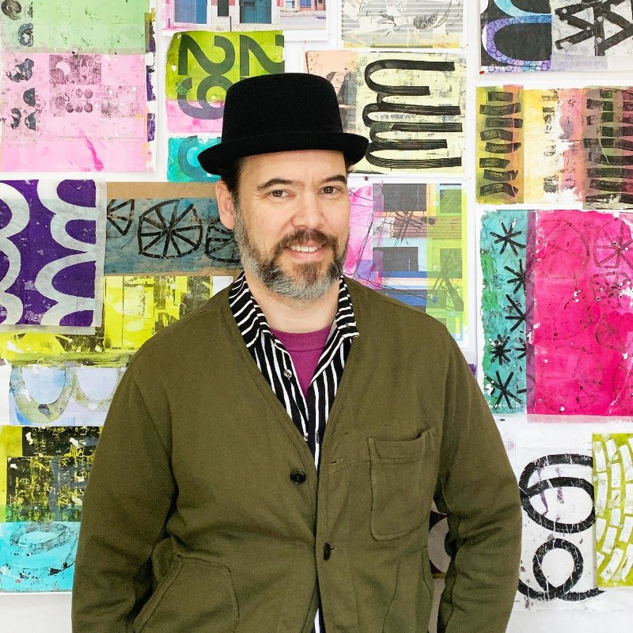 Smiling man in a black hat and olive jacket stands before a colorful wall of abstract prints and bold lettered art.