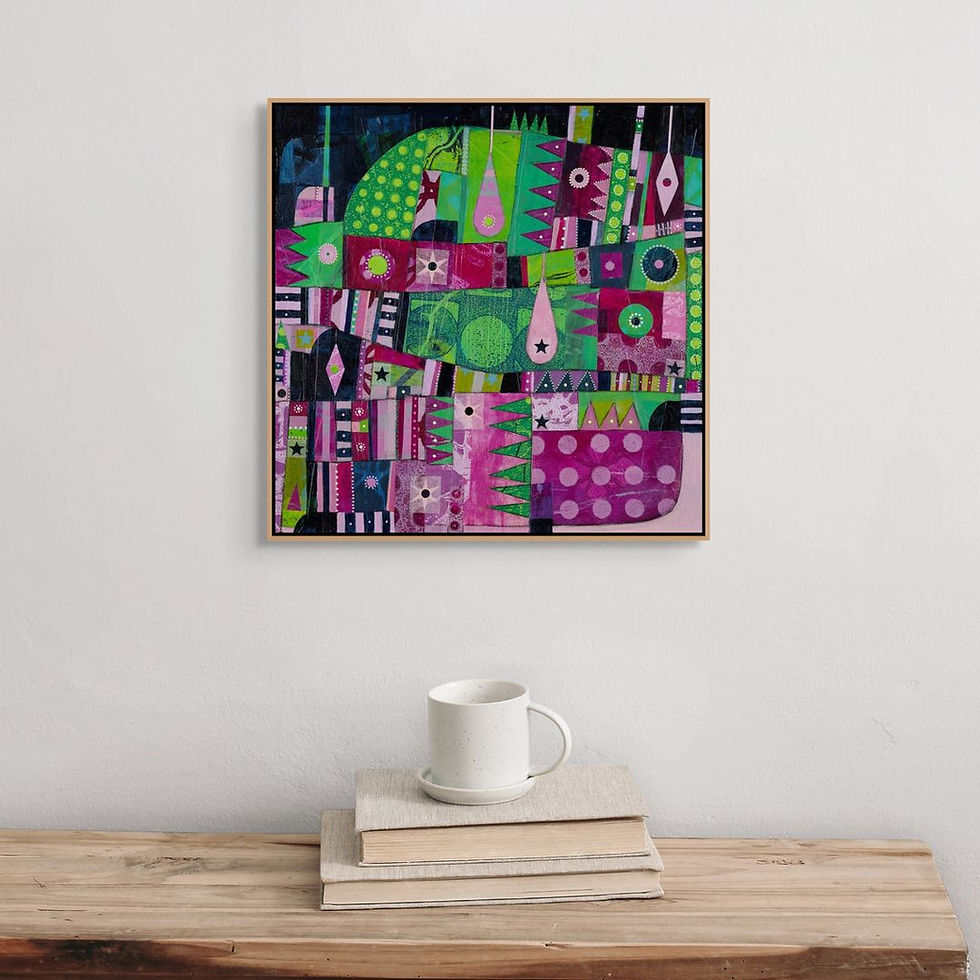 Colorful abstract painting with geometric shapes in pink, green, and blue. Framed on a white wall above a wooden table with books and a mug.