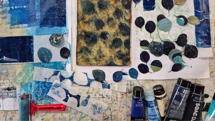 Art supplies, including paint tubes and a roller, on a colorful surface. Leaf prints and cutouts in blue and green tones are scattered on paper.