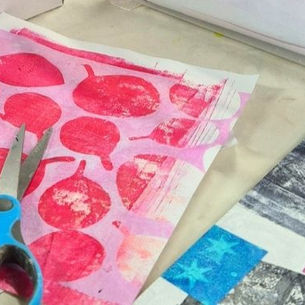 A table with colorful patterned papers, a blue-handled scissor, and a clear plastic bin. Abstract prints on wall in a creative studio.