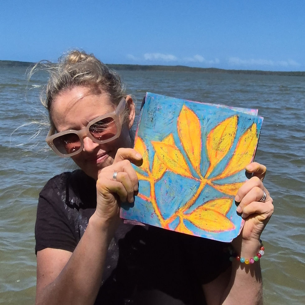 A person with sunglasses holds a colorful leaf painting by the ocean. The sky is clear, and the mood is cheerful.