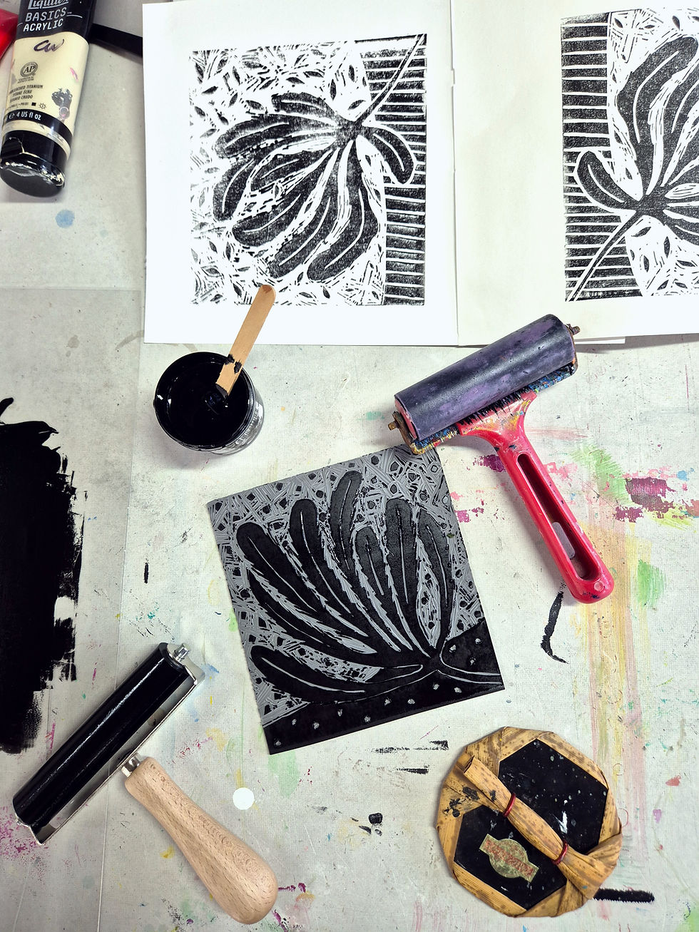 Printmaking setup with inked relief block, prints, rollers, and a black ink bottle on a messy, paint-splattered table.