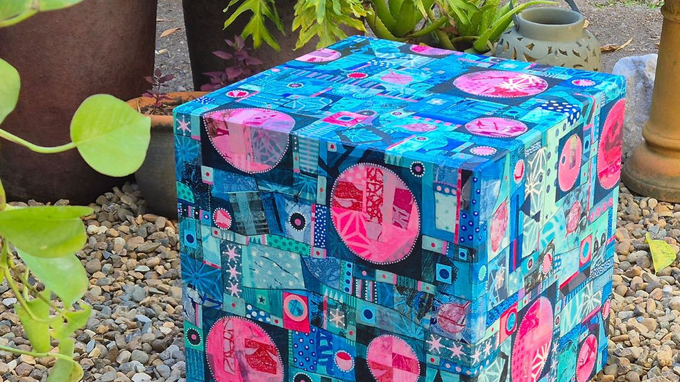 Colorful cube with blue and pink patterns sits on pebbles, surrounded by lush potted plants in a garden setting, creating a vibrant scene.