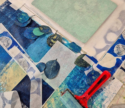 a gelli printin wrkspaceset up with scattered leaves, a gel plate, papers and a printmaking roller