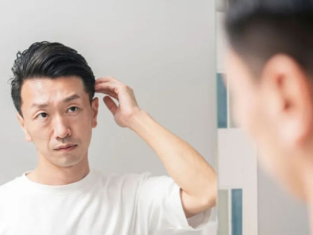 年輕男子在鏡子前整理頭髮,穿白色T恤,背景為淺灰色牆壁,表情專注。