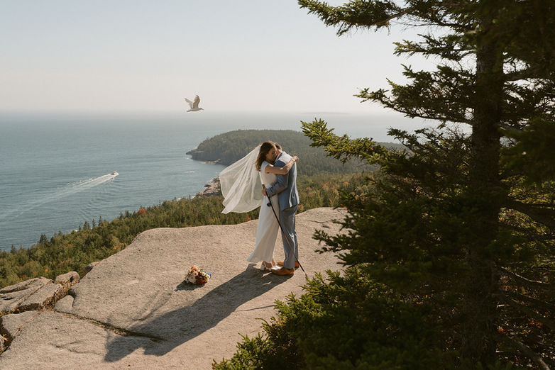 Couple sharing a hug on mountain top as a bird and boat go by in the background on wedding day in Acadia
