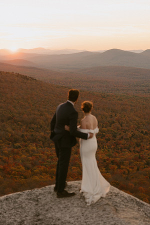 Couple exploring and enjoying sunset from a mountain top for Crawford Notch elopement day in the White Mountains for an adventure elopement. The mountains behind them are in peak foliage in nh and the sun is golden as it sets.