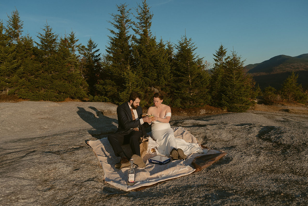 Couple exploring and enjoying sunset from a mountain top for Crawford Notch elopement day in the White Mountains for an adventure elopement. The mountains behind them are in peak foliage in nh and the sun is golden as it sets.