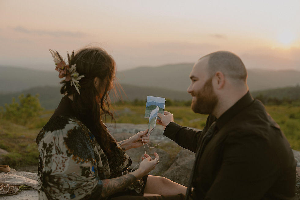 Couple holding vow books up to compare the view of the mountains with the mountains on vow books on wedding day on mountain top