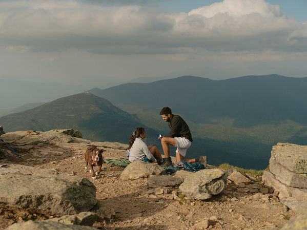Couple on Mount Lafayette summit, proposing with stunning view of white mountains in the background and dog by their side.